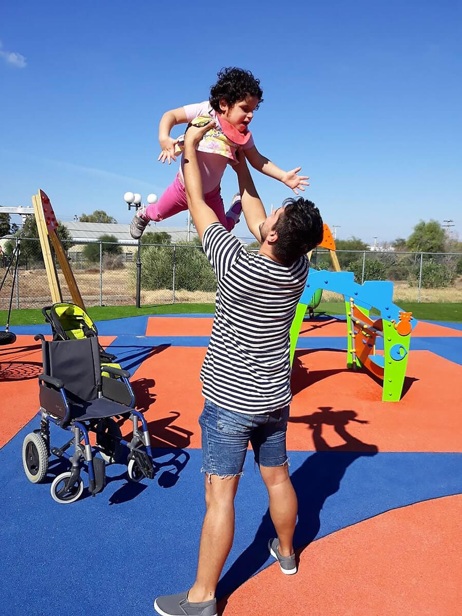amimoni.gr άνδρας κρατάει ψηλά ένα παιδάκι στην παιδική χαρά, αναπηρικό αμαξίδιο  | YouBeHero