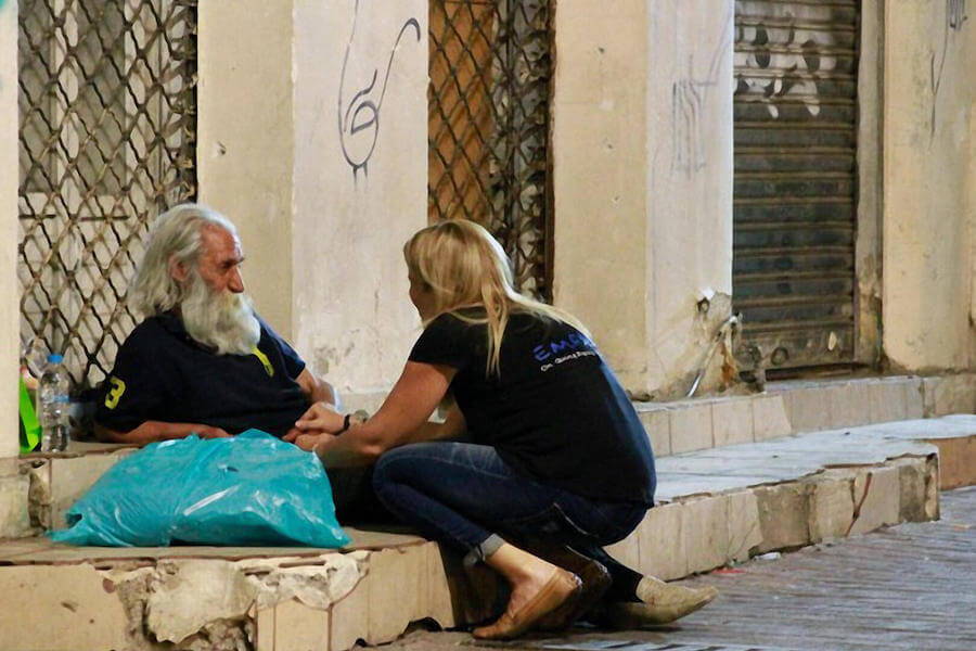 Εθελόντρια του emfasisfoundation.org προσεγγίζει άστεγο ξαπλωμένο στο πεζοδρόμιο με μία σακούλα γεμάτη ρούχα και είδη πρώτης ανάγκης | YouBeHero 